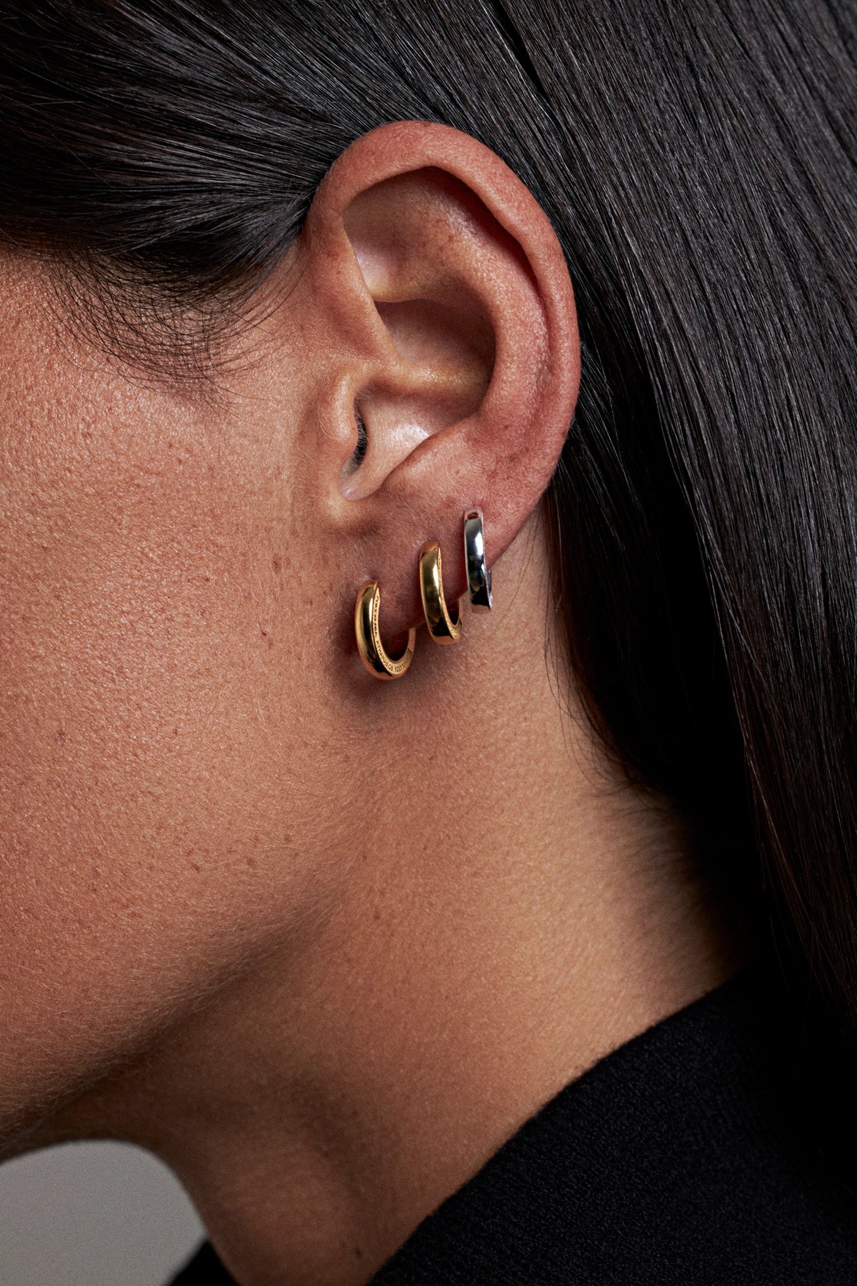 Close-up of a person's ear wearing three small hoop earrings in gold, silver, and black.