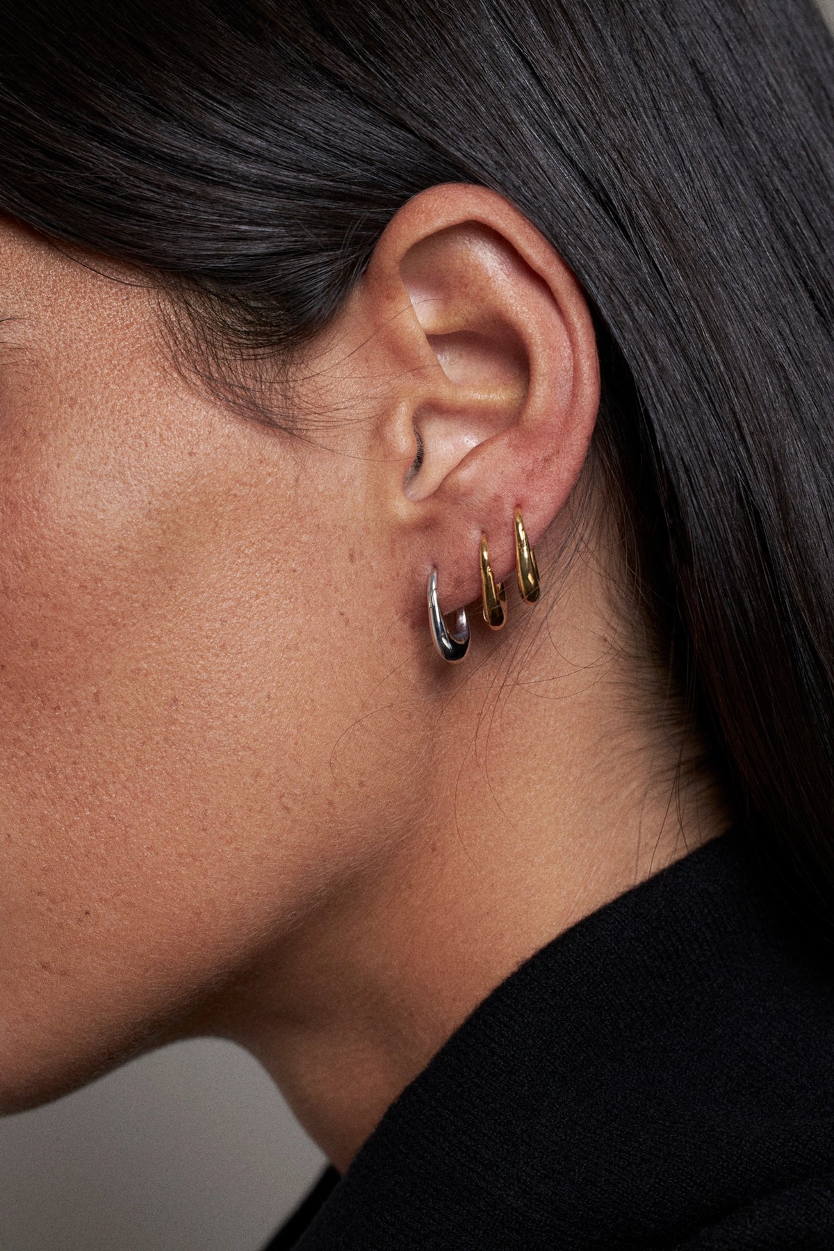 Close-up of a person's ear wearing three small hoop earrings in silver, gold, and rose gold tones, with dark hair and a black top visible.