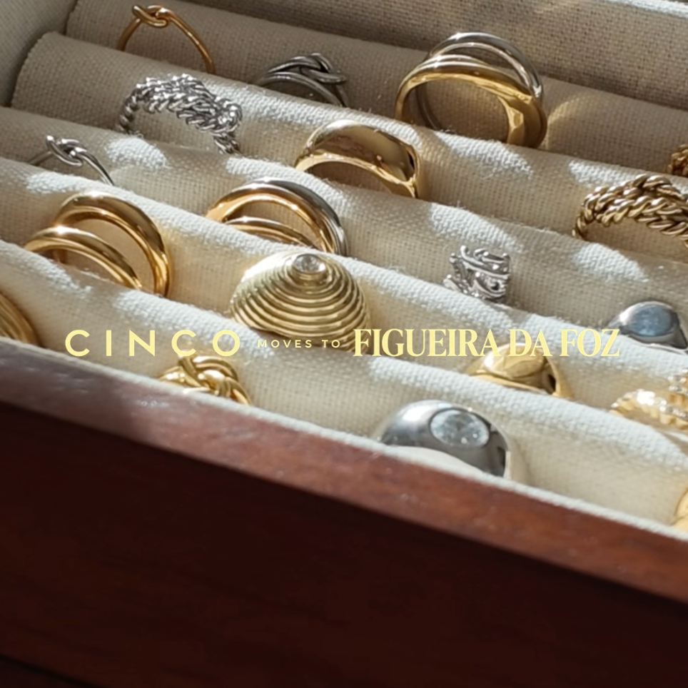 A close-up of a wooden jewelry box with cream fabric compartments holding multiple rings in gold and silver tones. A watermark reading 'CINCO LOVES FIGUEIRADATOZ' is visible across the center.