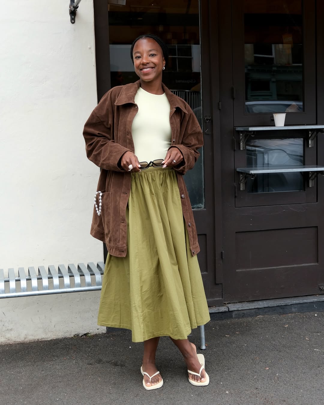 Smiling woman in a brown jacket and green skirt, standing outside a café with sunglasses in hand.
