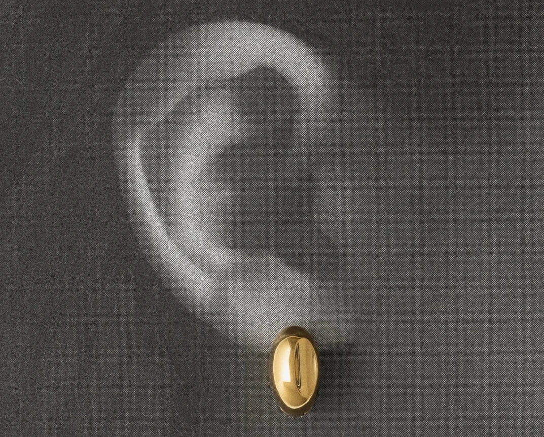 A small oval gold stud earring worn on a person's ear, shown against a dark charcoal gray textured background.