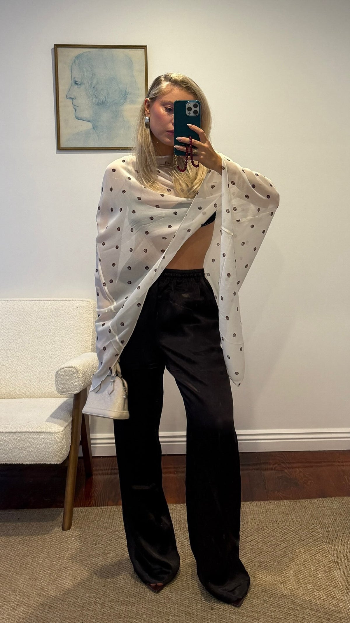 Woman wearing a white spotted shawl over a black outfit, holding a phone in front of a portrait, standing at home.