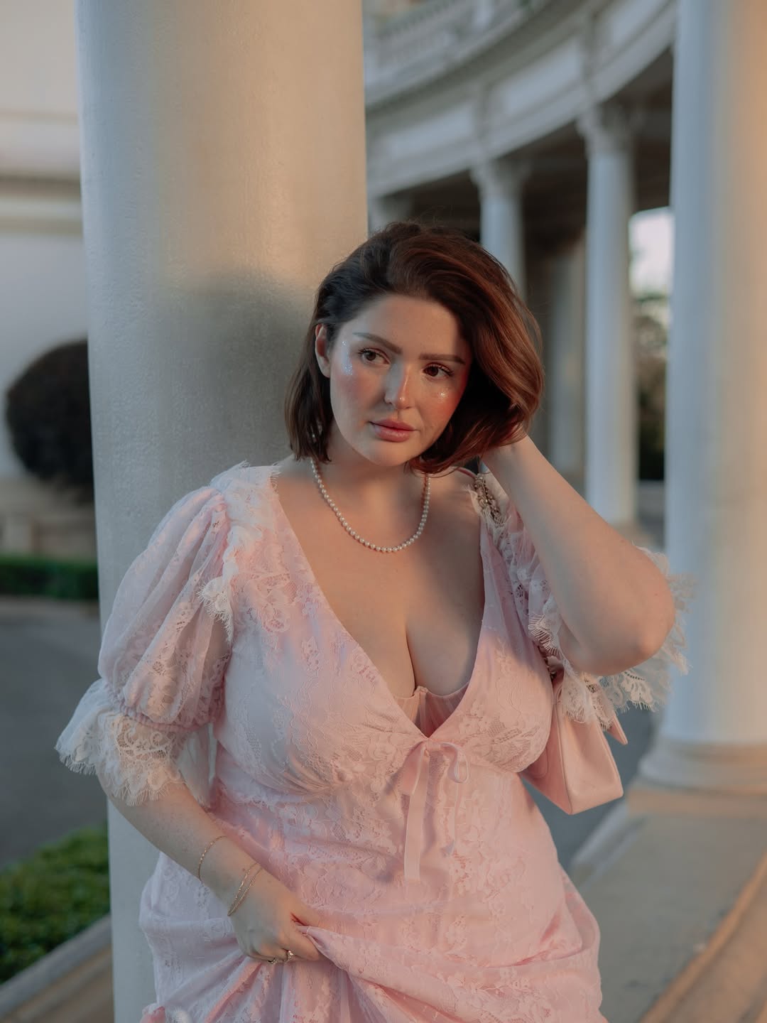 Woman in a pink lace dress with puff sleeves, wearing a pearl necklace, posing near a column.