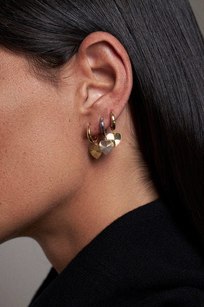 A close-up side profile of a person wearing a small gold hoop earring with dangling heart charms, one gold heart and one silver heart, against dark hair and a black top.