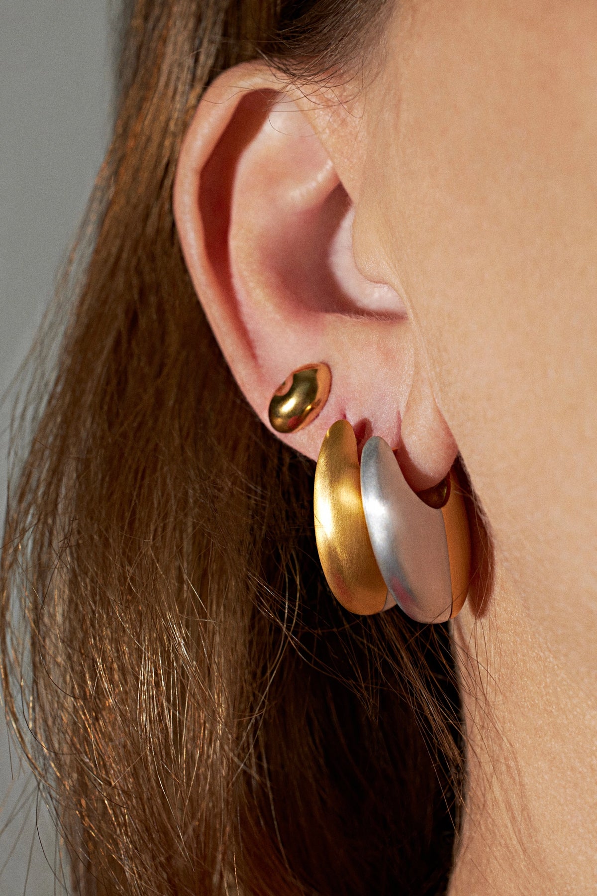 Close-up of a model's ear wearing a stack of three crescent hoop earrings in gold, rose gold, and silver tones, with brown hair framing the side profile.
