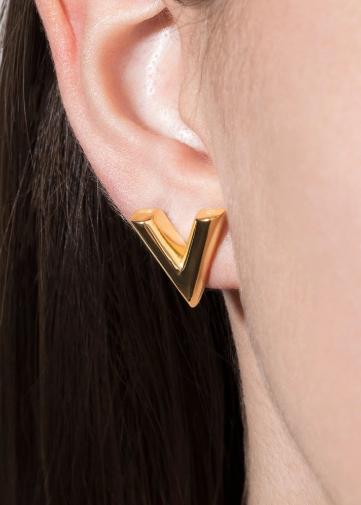 A close-up of a person's ear wearing a gold geometric letter V stud earring.